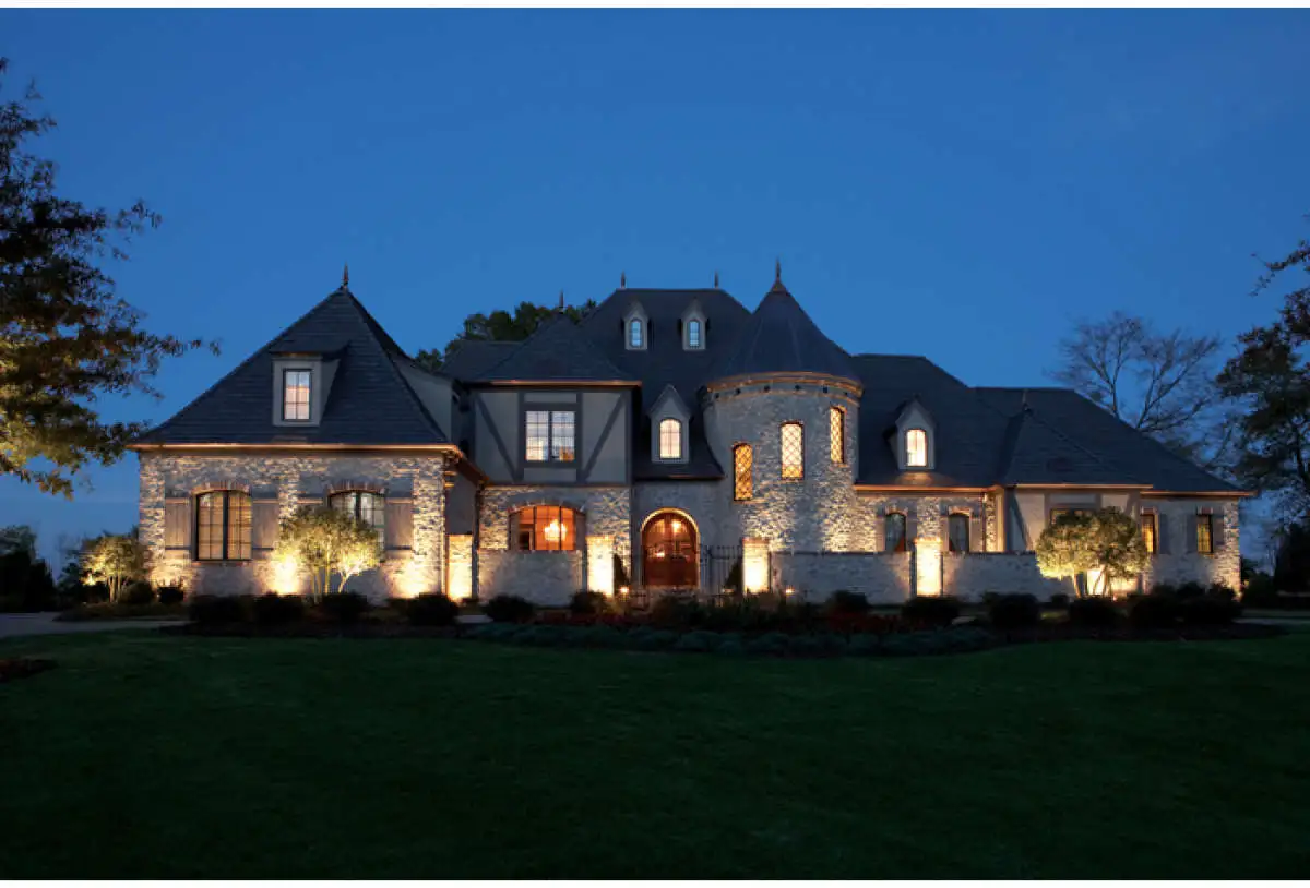 7-bedroom-house-plan.webp Large stone house with turrets and steep roofs, exterior lights illuminating the building and surrounding landscaping at dusk—perfect for adding to your house Plan Wishlist.
