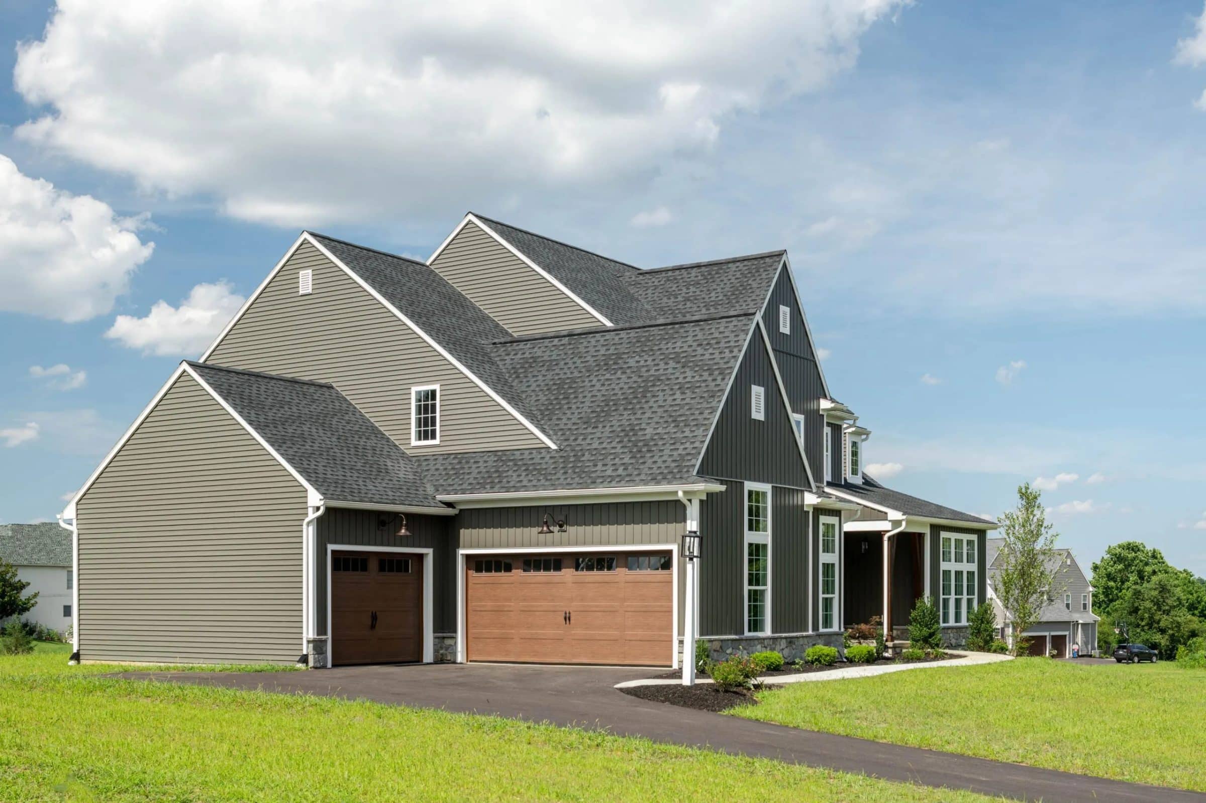 A modern two-story house with gray siding, a three-car garage with wooden doors, and a paved driveway sits on a grassy lawn under a partly cloudy sky—perfect for adding to your house Plan Wishlist.